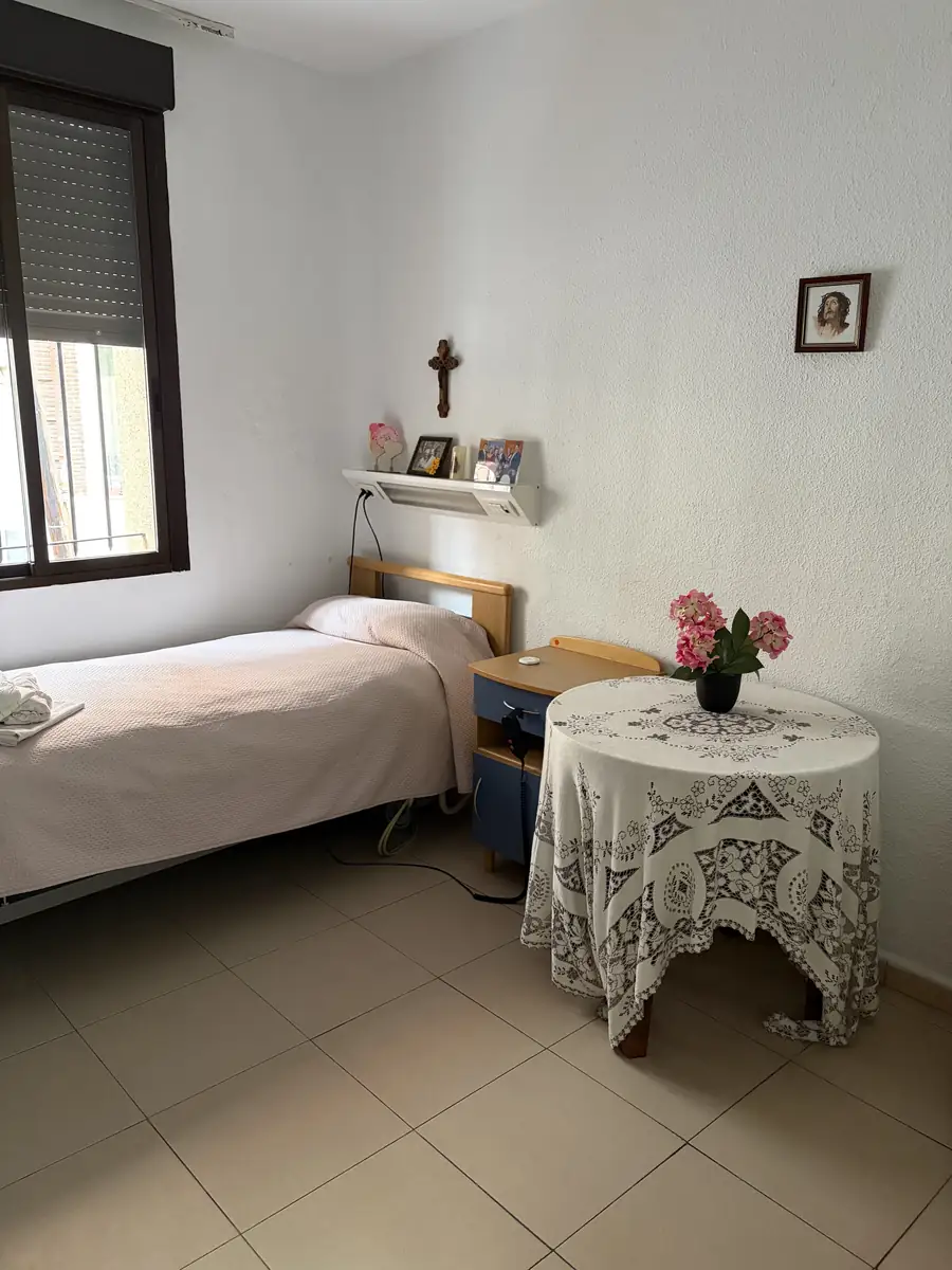 Habitación con cama, mesa camilla con flores y ventana