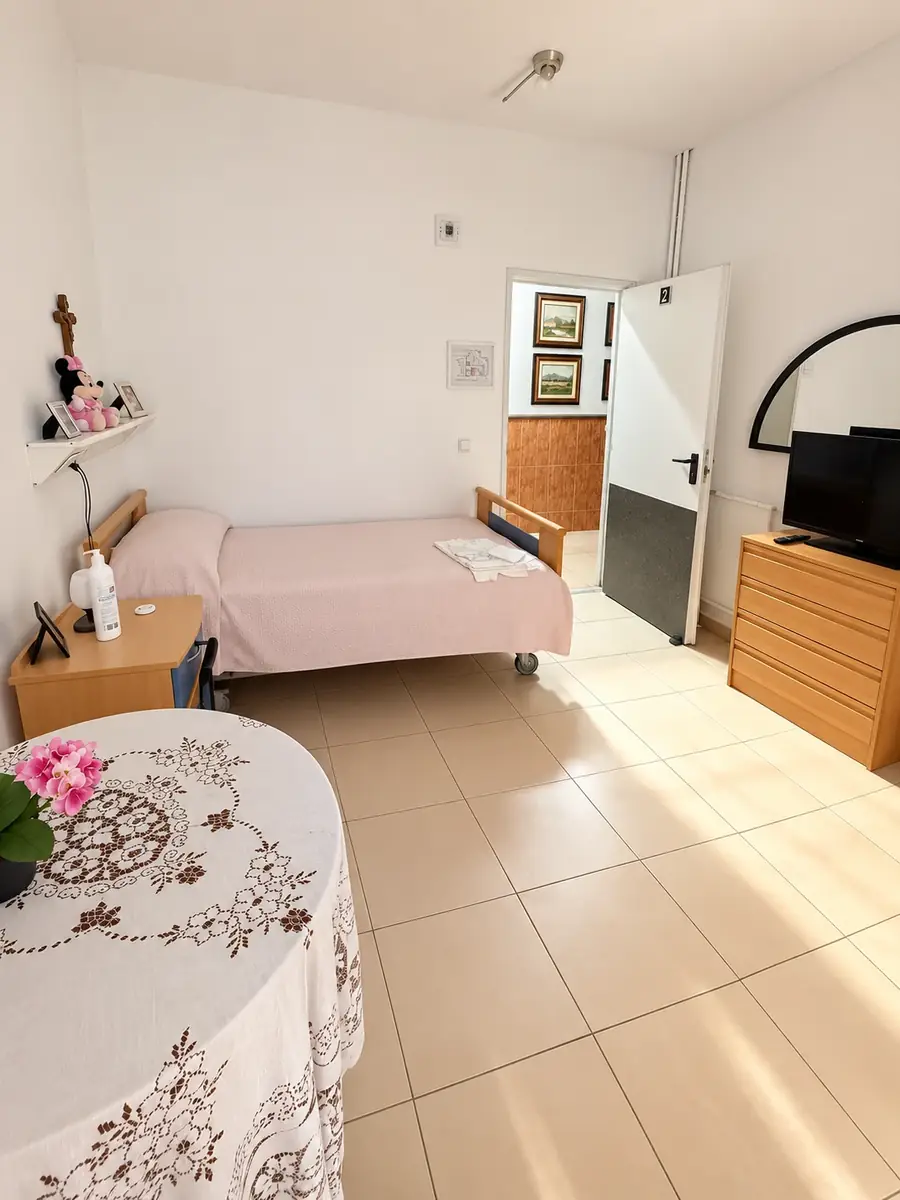 Habitación luminosa con cama, mesa camilla con flores, cómoda y televisión